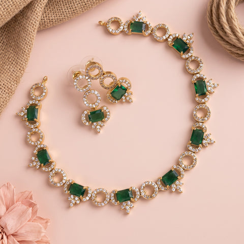 Gold-plated jewelry set featuring a necklace, matching earrings, and a bracelet, decorated with green rectangular gemstones and clear rhinestones, arranged on a light pink background with decorative burlap and a flower.