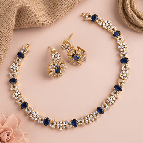 Gold-tone jewelry set featuring a necklace and matching earrings with clear crystal floral clusters and dark blue oval accents, arranged on a soft pink background with burlap and a rose prop.