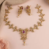 Gold-toned floral jewelry set with a matching necklace and earrings, featuring clear rhinestones and small purple teardrop gemstones, arranged on a soft pink background with a beige fabric corner and a pale pink flower.