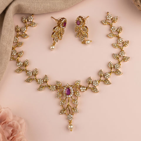 Gold-toned floral jewelry set with a matching necklace and earrings, featuring clear rhinestones and small purple teardrop gemstones, arranged on a soft pink background with a beige fabric corner and a pale pink flower.