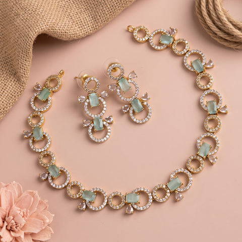 Gold-tone jewelry set with circular crystal-studded rings and pale green rectangular stones, including a necklace, matching earrings, and a bracelet, arranged on a soft pink background with burlap and a pink flower prop.