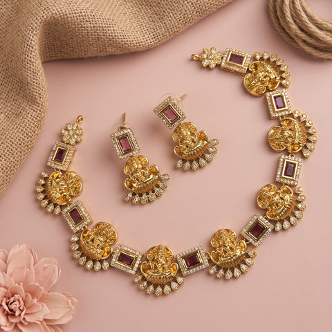 Gold-toned traditional jewelry set with Ganesha motifs, rectangular maroon stone accents, and clear crystal embellishments, arranged on a pink background with burlap fabric and a pink flower.