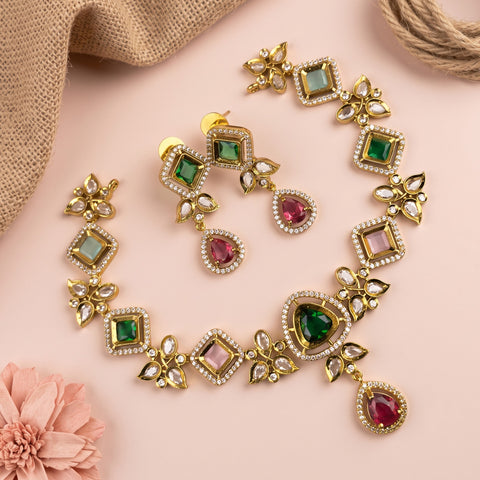 Gold-plated jewelry set featuring a necklace and matching earrings with green, pink, and light blue stones, displayed on a soft pink background with decorative burlap and a flower.