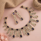 Gold-toned traditional jewelry set with a necklace, earrings, and bracelet, featuring purple teardrop gemstones and clear rhinestone-like accents, arranged on a pink surface with burlap fabric and a flower.