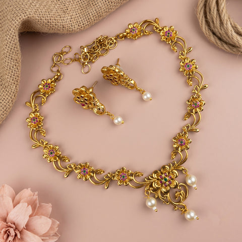 Gold-toned floral jewelry set with a matching necklace and earrings, featuring small pink gemstone accents and dangling white faux pearls, arranged on a soft pink background with burlap and a pink flower prop.