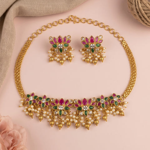 Gold-toned traditional necklace and matching earrings set with floral motifs, featuring pink stones, green stones, and dangling pearl-like beads, arranged on a soft pink background with decorative fabric and a flower prop.