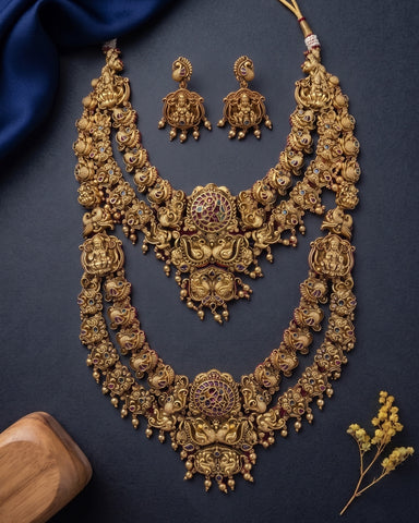 An ornate gold-toned traditional necklace set with matching earrings, displayed on a dark navy fabric background with decorative blue satin and small dried flowers.