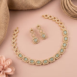 Gold-plated jewelry set featuring a choker necklace and matching drop earrings with rectangular light green stones surrounded by small clear crystals, arranged on a pink background with burlap and a flower prop.