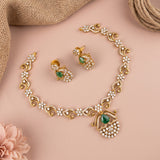 Gold-toned traditional jewelry set with a necklace, earrings, and matching earrings, decorated with white stones and green teardrop gemstones, arranged on a pink background with burlap and a pink flower.