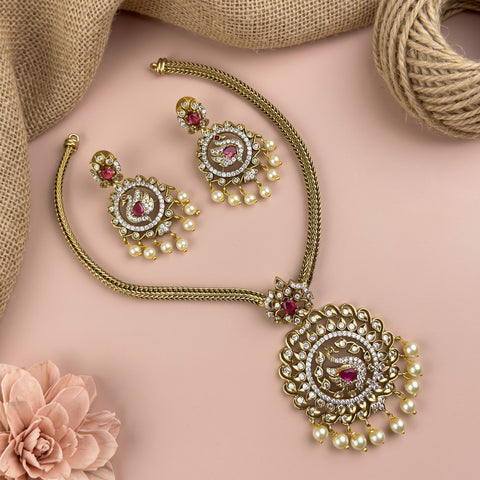 Gold-toned Indian traditional jewelry set featuring a necklace and matching earrings with intricate filigree, white faux pearls, and pink gemstone accents, arranged on a soft pink background with burlap and a flower prop.