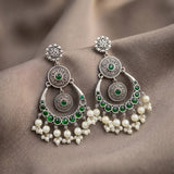 A pair of ornate silver-toned dangle earrings with green stones and white pearl-like beads, displayed on a draped taupe fabric background.