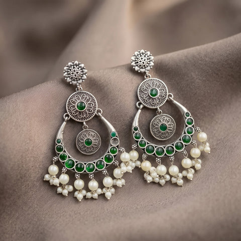 A pair of ornate silver-toned dangle earrings with green stones and white pearl-like beads, displayed on a draped taupe fabric background.