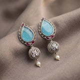 A pair of ornate silver-toned dangle earrings with light blue teardrop-shaped stones, small red gem accents, striped metal beads, clear rhinestones, and pearl drops, displayed on draped taupe fabric.
