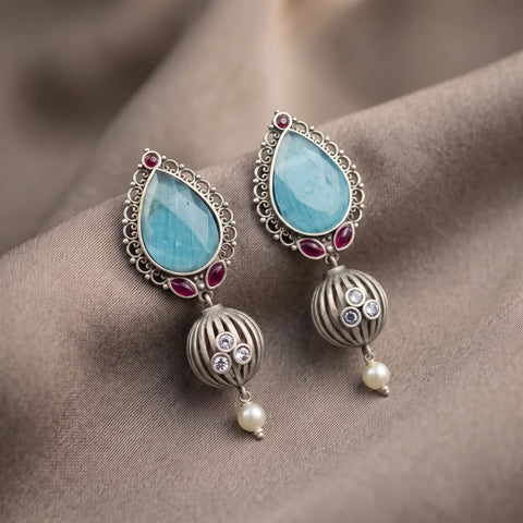A pair of ornate silver-toned dangle earrings with light blue teardrop-shaped stones, small red gem accents, striped metal beads, clear rhinestones, and pearl drops, displayed on draped taupe fabric.