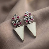 A pair of silver-toned geometric dangle earrings with a floral cluster top featuring red and light blue stones, and a large white triangular drop below, displayed on draped taupe fabric.