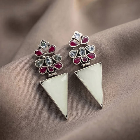 A pair of silver-toned geometric dangle earrings with a floral cluster top featuring red and light blue stones, and a large white triangular drop below, displayed on draped taupe fabric.