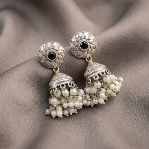 A pair of silver-toned Indian jhumka earrings with black stone centers and dangling white pearl-like beads, displayed on a draped taupe fabric background.