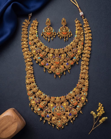 An ornate gold-toned traditional jewelry set laid flat on a dark navy background, including a long necklace, matching earrings, and a smaller coordinating piece. The jewelry is heavily decorated with colorful enamel-like accents in orange, blue, and red, with small dangling beads.