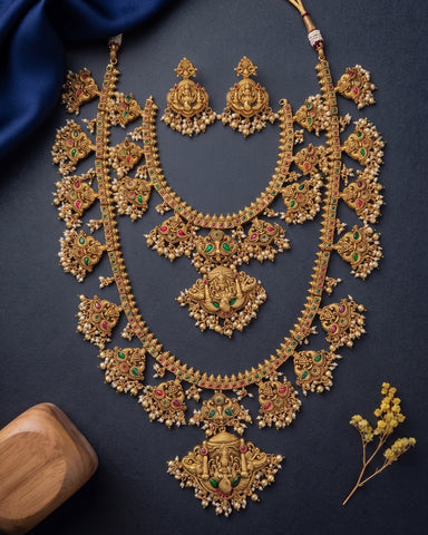 An ornate gold-toned traditional necklace set with matching earrings, decorated with intricate filigree, beadwork, and red and green stone accents, arranged on a dark navy background with a blue fabric corner and small floral props.