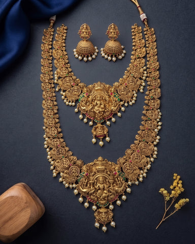 An ornate gold-toned traditional jewelry set laid flat on a dark navy background, including a layered necklace with a large central deity pendant, matching earrings, and pearl bead accents. A blue fabric drape, wooden block, and small dried floral sprig are placed around the set as styling props.