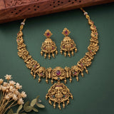 An ornate gold-toned traditional necklace and matching earrings set with red and green gemstone accents, arranged on a dark green background with decorative dried flowers and a carved wooden prop.