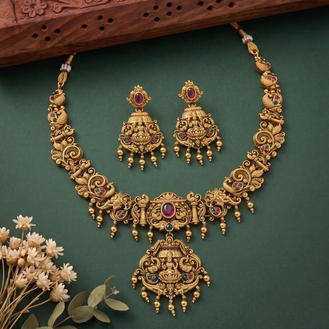 An ornate gold-toned traditional necklace and matching earrings set with red and green gemstone accents, arranged on a dark green background with decorative dried flowers and a carved wooden prop.