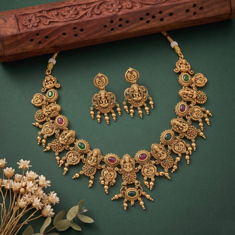 An ornate gold-toned traditional necklace and matching earrings set with green and red stone accents, arranged on a dark green background with a carved wooden prop and small dried flowers.