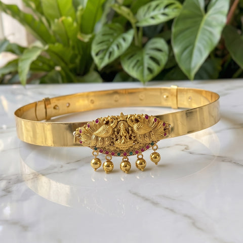 Gold-plated metal bangle bracelet with a detailed central ornament featuring peacock motifs and hanging gold beads, displayed on a white marble surface with green plants in the background.