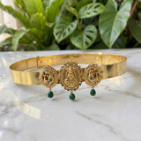 Gold-toned traditional Indian jewelry set featuring a bangle and matching earrings with ornate deity motifs, green bead drops, and small red and green stone accents, displayed on a white marble surface with green foliage in the background.
