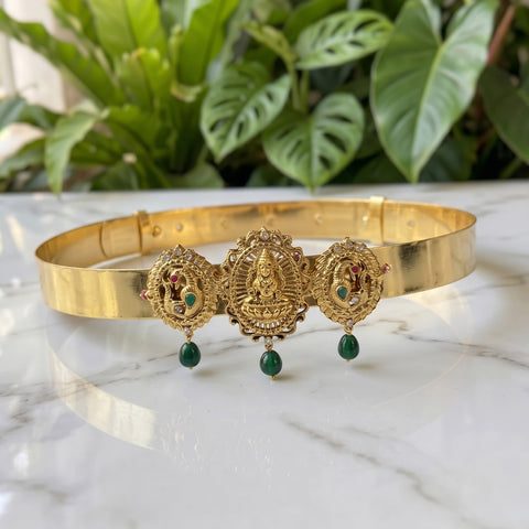 Gold-toned traditional Indian jewelry set featuring a bangle and matching earrings with ornate deity motifs, green bead drops, and small red and green stone accents, displayed on a white marble surface with green foliage in the background.