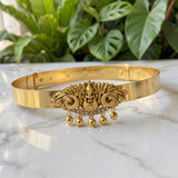A gold-toned traditional Indian choker belt featuring an ornate central deity motif, small rhinestone accents, and dangling bell charms, displayed on a white marble surface with green foliage in the background.