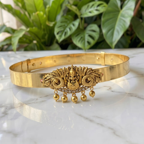 A gold-toned traditional Indian choker belt featuring an ornate central deity motif, small rhinestone accents, and dangling bell charms, displayed on a white marble surface with green foliage in the background.