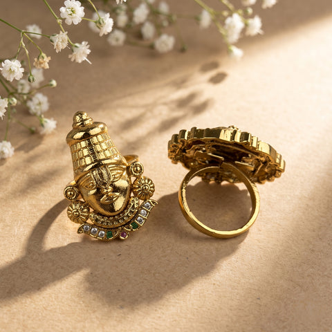 A pair of gold-toned statement rings displayed on a textured beige surface with soft shadows and small white flowers. One ring is shown from the front, featuring a detailed deity face with a crown and small multicolor gem accents.