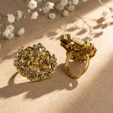 Two ornate gold rings displayed on a textured beige surface with soft shadows and small white flowers in the background. One ring features an intricate deity motif with small green and red stones, and the other is a polished gold ring.