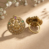 An ornate gold-toned traditional Indian jewelry piece displayed in two views on a beige textured surface with soft shadows and small white flowers in the background. The piece features a detailed central motif with green and purple stones and a circular ring attached.