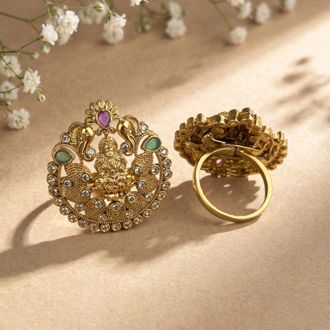 An ornate gold-toned traditional Indian jewelry piece displayed in two views on a beige textured surface with soft shadows and small white flowers in the background. The piece features a detailed central motif with green and purple stones and a circular ring attached.