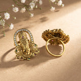 Two ornate gold rings displayed on a beige surface with soft shadows and small white flowers in the background. One ring features a detailed deity face with a halo of clear stones and green gem accents, while the other shows the textured back of the band.