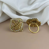 Gold ring with intricate design and gemstones on a beige surface with white fabric background