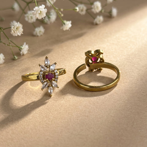Two gold rings displayed on a beige surface with soft shadows and small white flowers in the background. One ring features a starburst cluster with clear marquise-cut stones and a central round pink gemstone, while the other shows a side view of a matching pink gemstone setting.