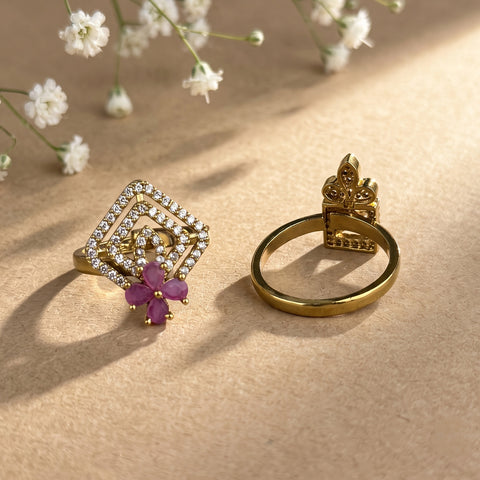 Two gold rings displayed on a textured beige surface with soft shadows and small white flowers in the background. One ring features a square pavé setting with clear stones and a purple flower accent, and the other shows the back of a gold band with a decorative crest-like top.