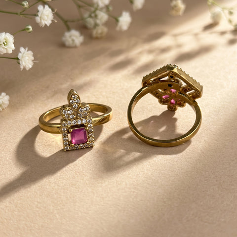 Two gold rings with pink gemstones and clear stones, styled on a beige surface with soft shadows and small white flowers.