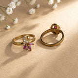 Two gold rings displayed on a beige surface with soft shadows and small white flowers in the background. One ring has a butterfly-shaped setting with clear stones and pink marquise-cut stones, and the other shows the back with a round pink stone.