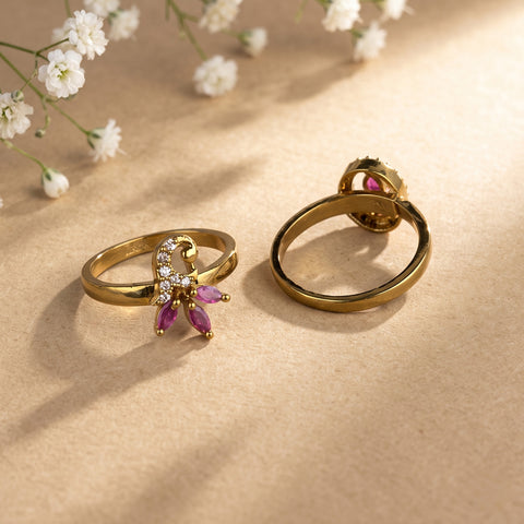 Two gold rings displayed on a beige surface with soft shadows and small white flowers in the background. One ring has a butterfly-shaped setting with clear stones and pink marquise-cut stones, and the other shows the back with a round pink stone.