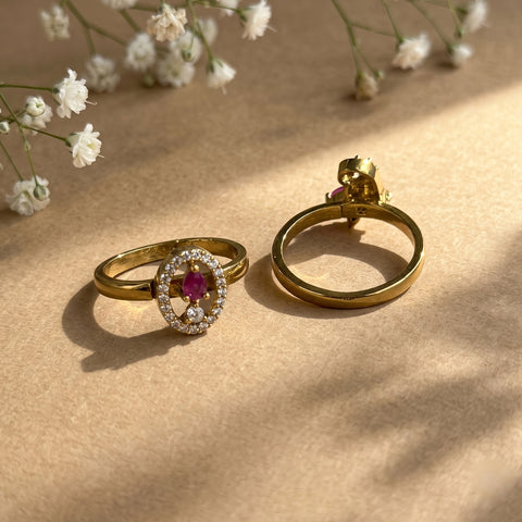 A pair of gold rings displayed on a textured beige surface with soft shadows and small white flowers in the background. One ring features a round halo setting with a central pink gemstone and clear stones around it, while the other shows the back of the gold band.