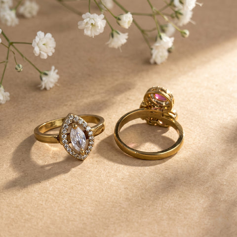 Two gold rings displayed on a textured beige surface with soft shadows and small white flowers in the background. One ring features a marquise-shaped clear gemstone surrounded by small clear stones, and the other shows a gold band with a decorative clasp and a pink gemstone.