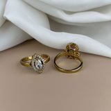Two gold rings with gemstones on a beige surface with white fabric in the background