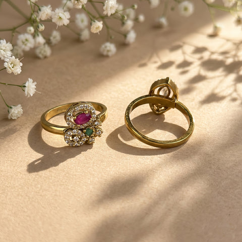 Two gold rings displayed on a beige surface with soft shadows and small white flowers. One ring is shown from the front with a floral cluster setting featuring a pinkish-red oval center stone, small clear stones, and a tiny green accent.