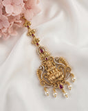 An ornate gold-toned traditional jewelry piece with red gemstone accents and white pearl drops, styled on a draped white fabric with pink flowers in the corner.