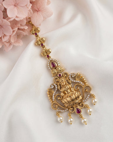 An ornate gold-toned traditional jewelry piece with red gemstone accents and white pearl drops, styled on a draped white fabric with pink flowers in the corner.