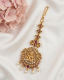 An ornate gold-toned traditional Indian jhumka earring decorated with red gemstones, white pearls, and a central deity motif, displayed on a white satin fabric next to pink flowers.